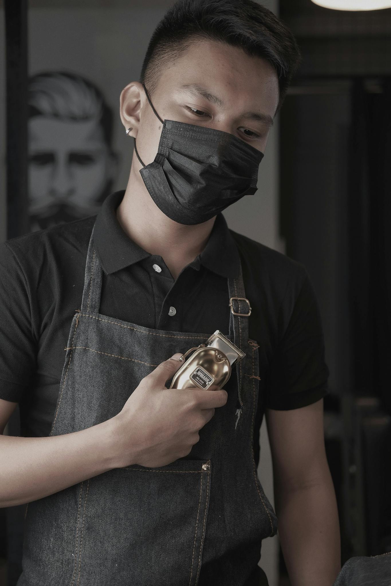 Barber in apron and mask holding hair clipper inside a salon, ready for work.
